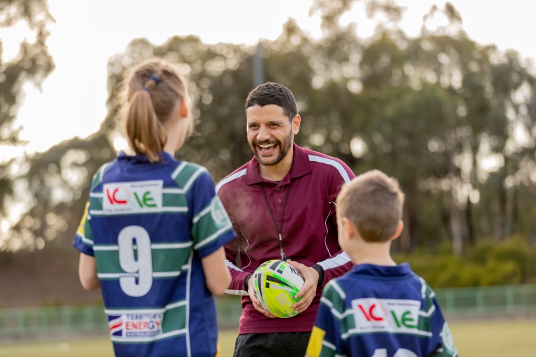 A coach holding a rugby ball smiles at two junior players