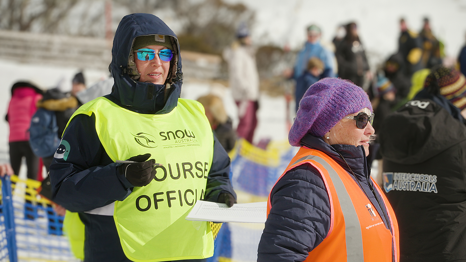 A course official with a snow jacket and yellow bib with 'course official' holds a piece of paper and watches the competition. 