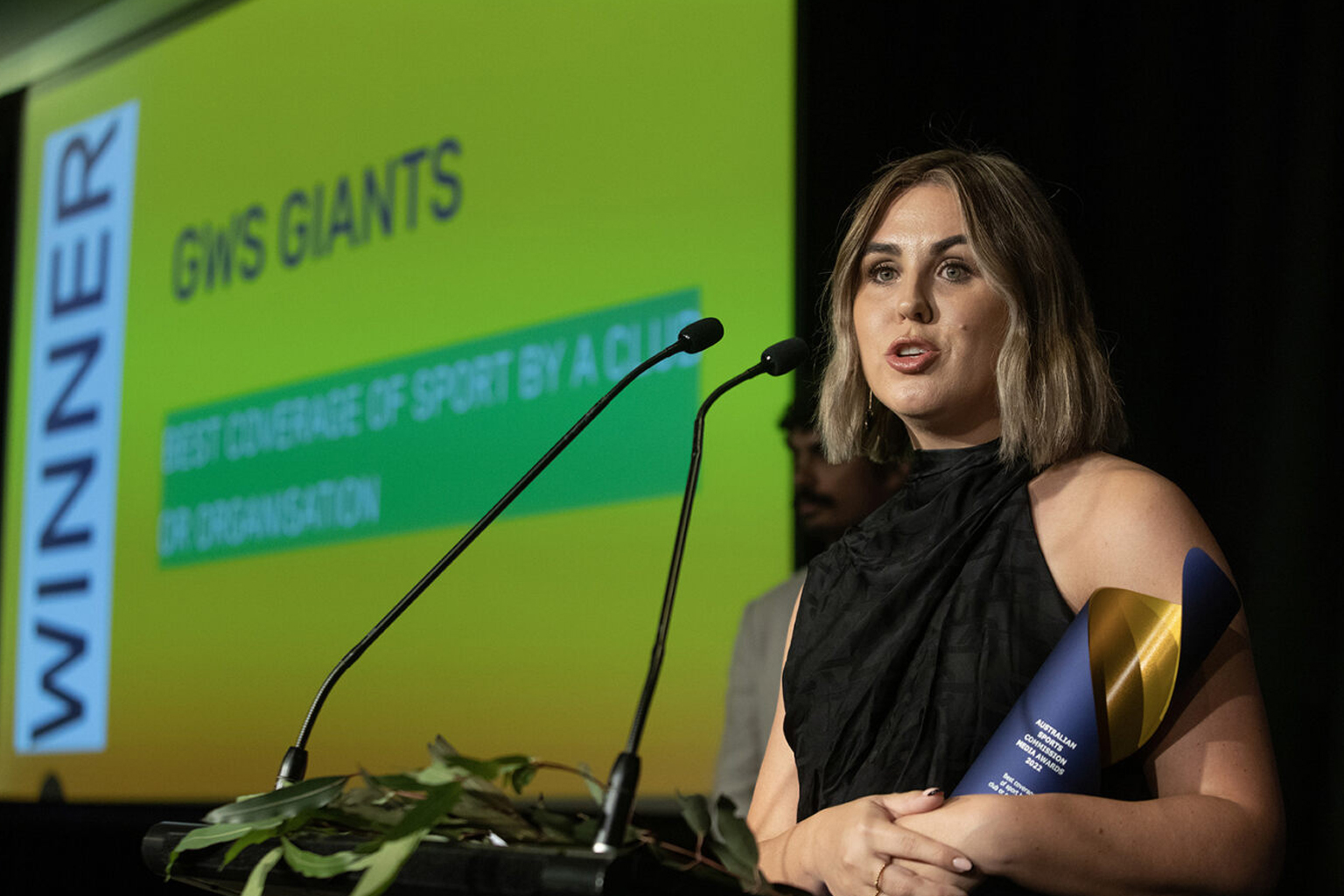 A woman stands at a lectern holding a trophy with Winner GWS Giants written on a screen in the background